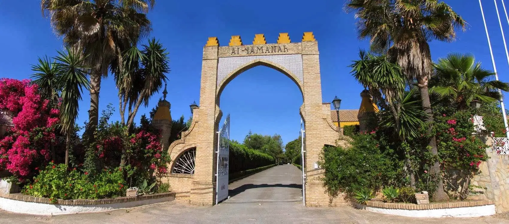 El arco de entrada almenado, con el nombre Al-Yamanah, entre palmeras y buganvillas