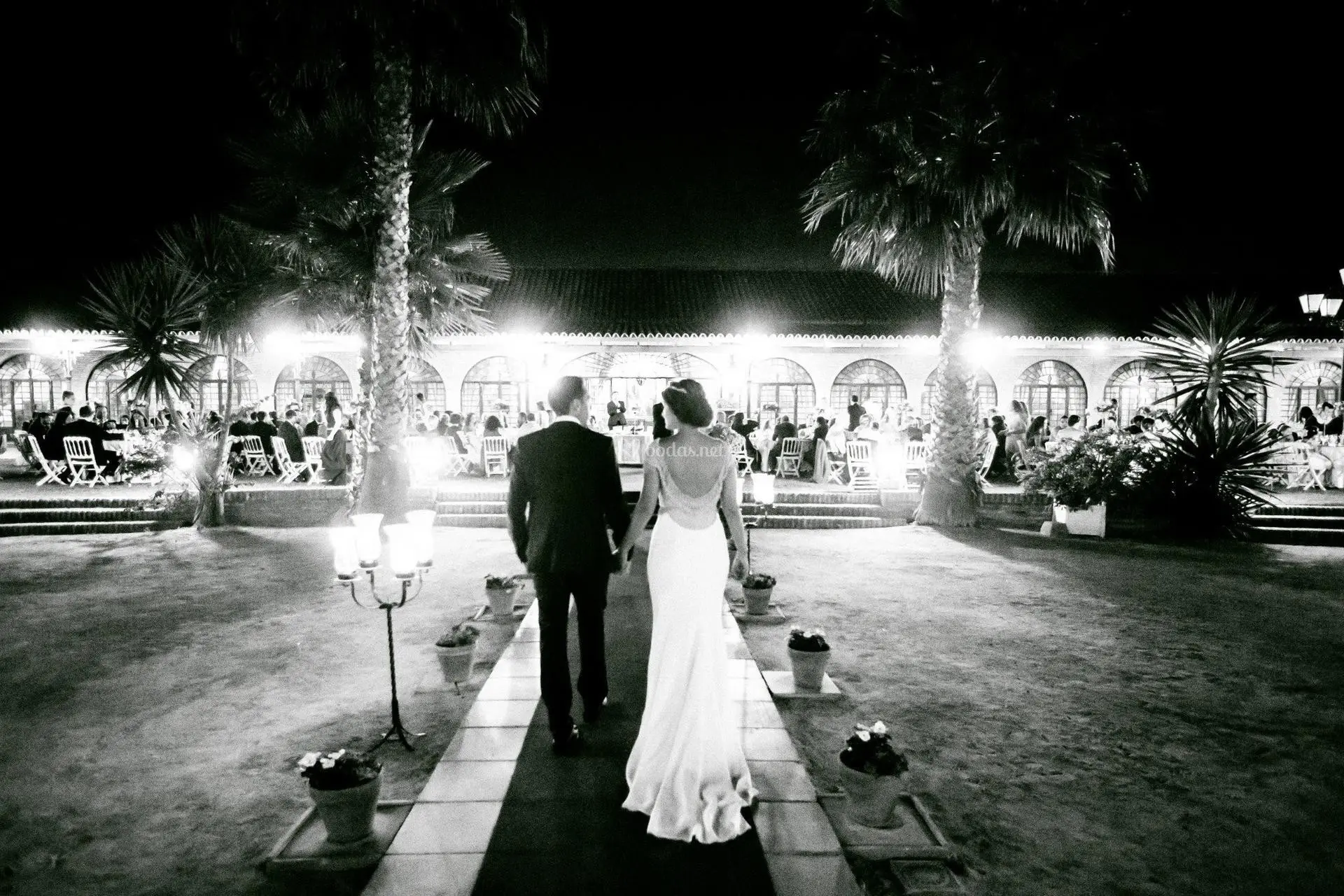 Los novios, de espaldas, caminan de noche hacia la arcada iluminada