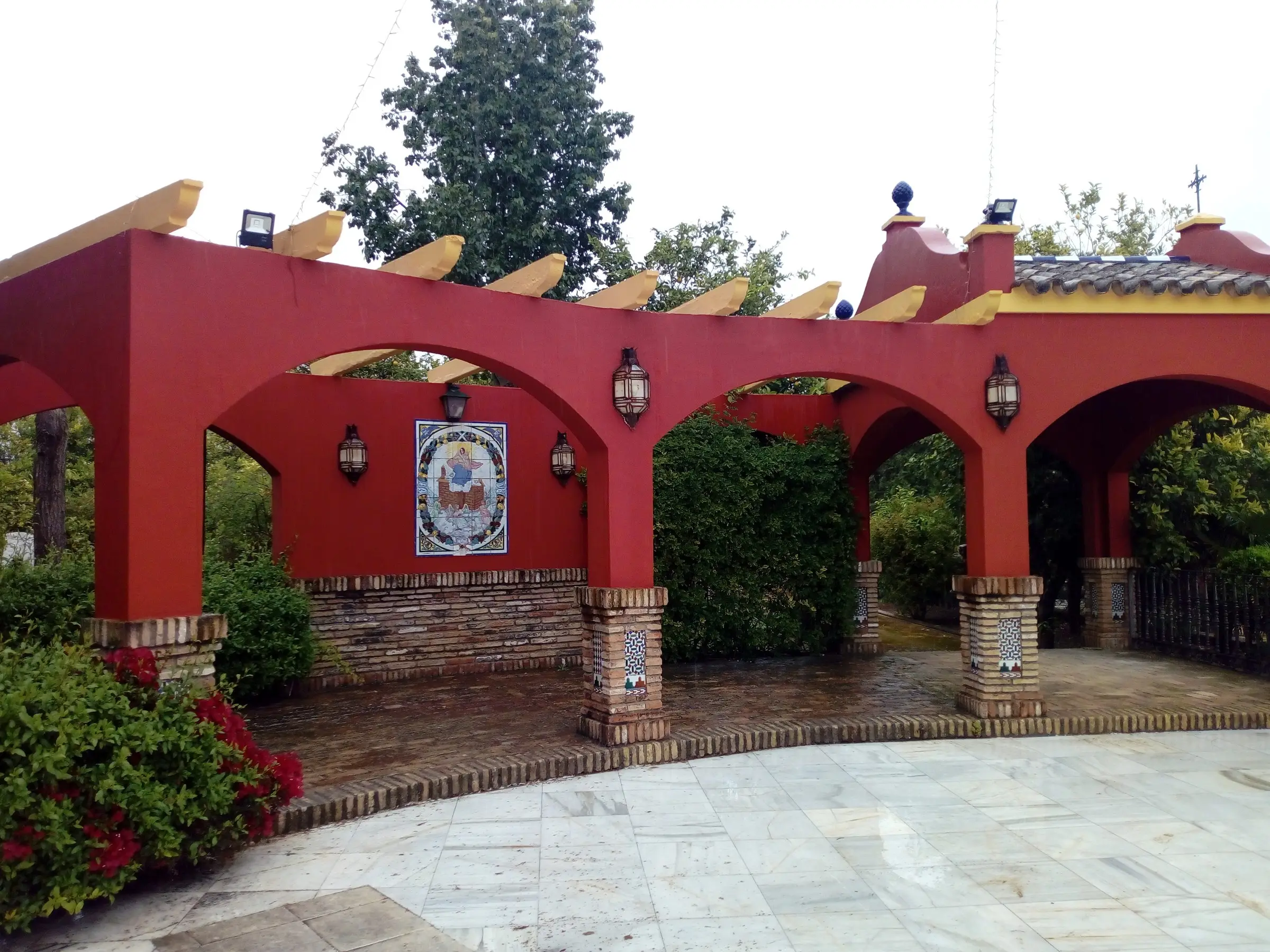 Pabellón de arcos rojos con un retablo cerámico y lámparas de forja