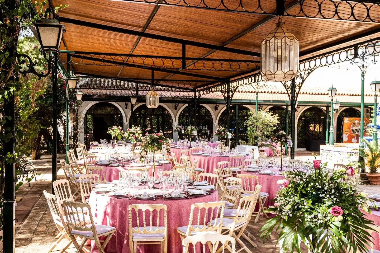 Mesas con mantel rosa bajo una pérgola de forja con farolillos