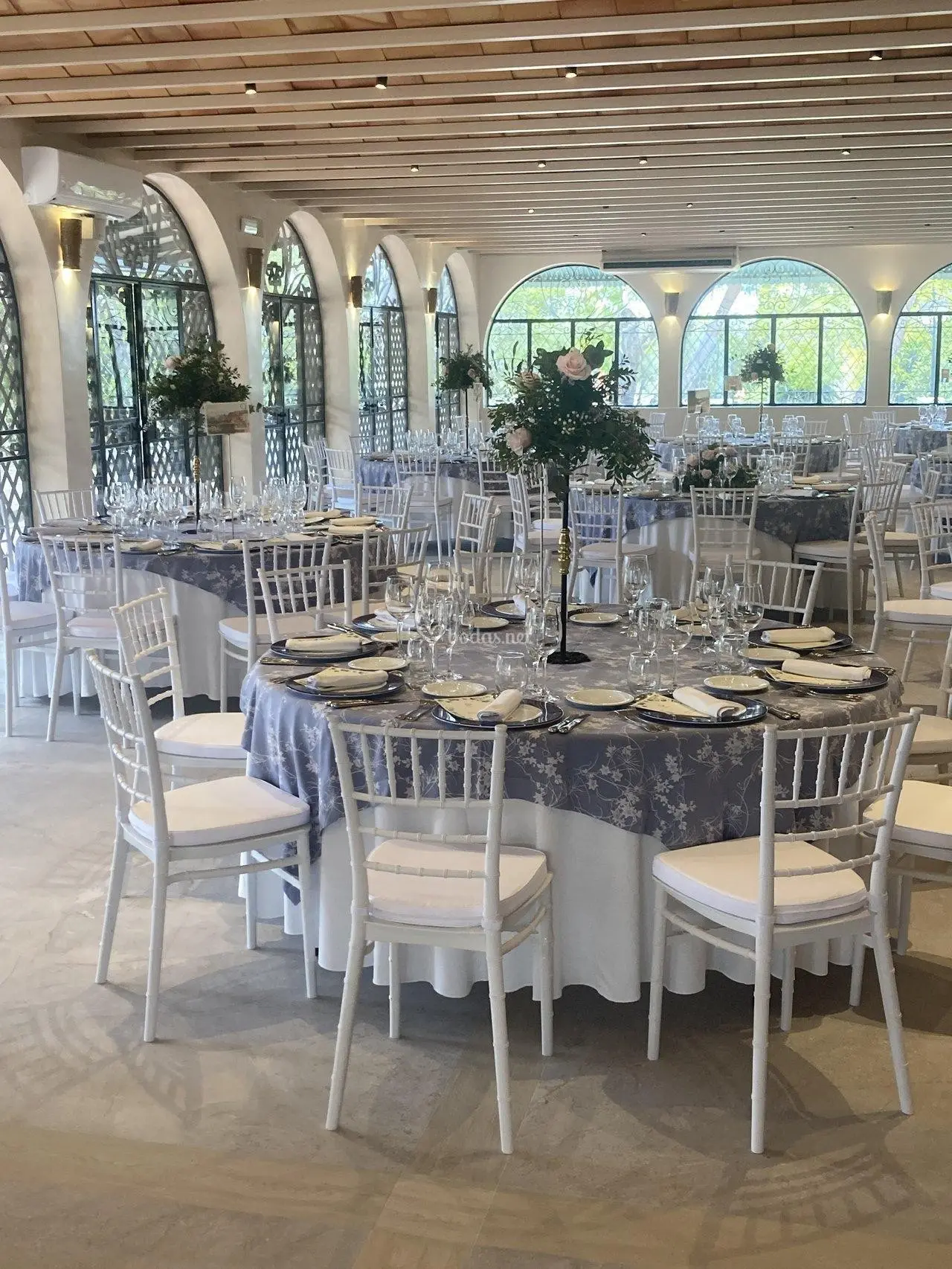Salón blanco con ventanales en arco, mesas vestidas y sillas blancas